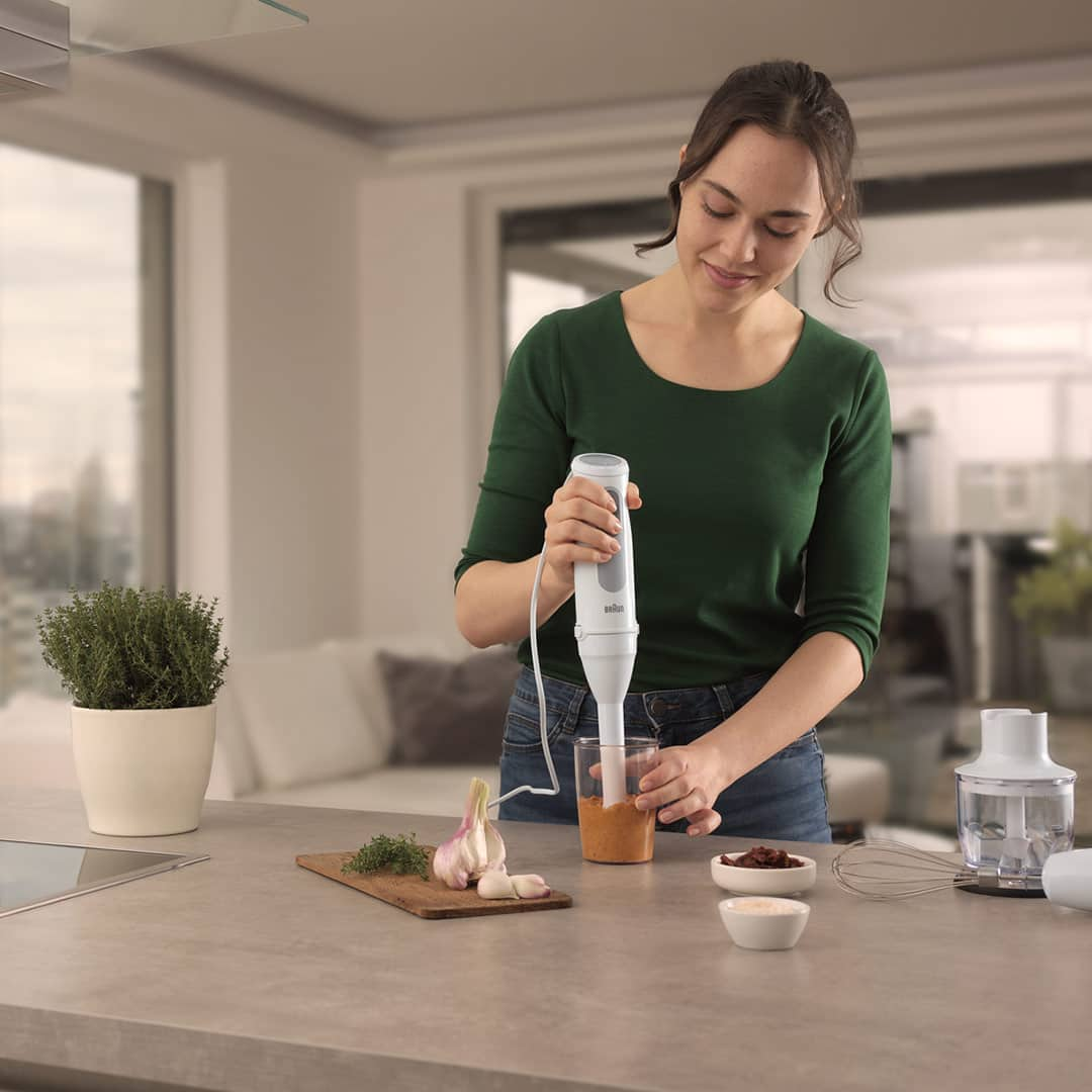 Woman at the kitchen counter preparing a vegetable mash with the Braun MultiQuick 5 with Small Portion Shaft accessory. Small chopper and whisk attachment are laying aside on the desk.