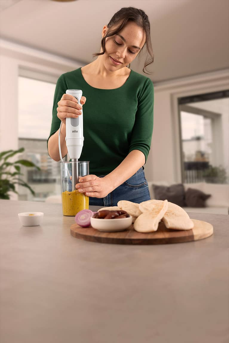 A young woman at a kitchen counter preparing a dip with the MultiQuick 5 Hand blender. In the foreground, a plate with pita bread, dates and half an onion.