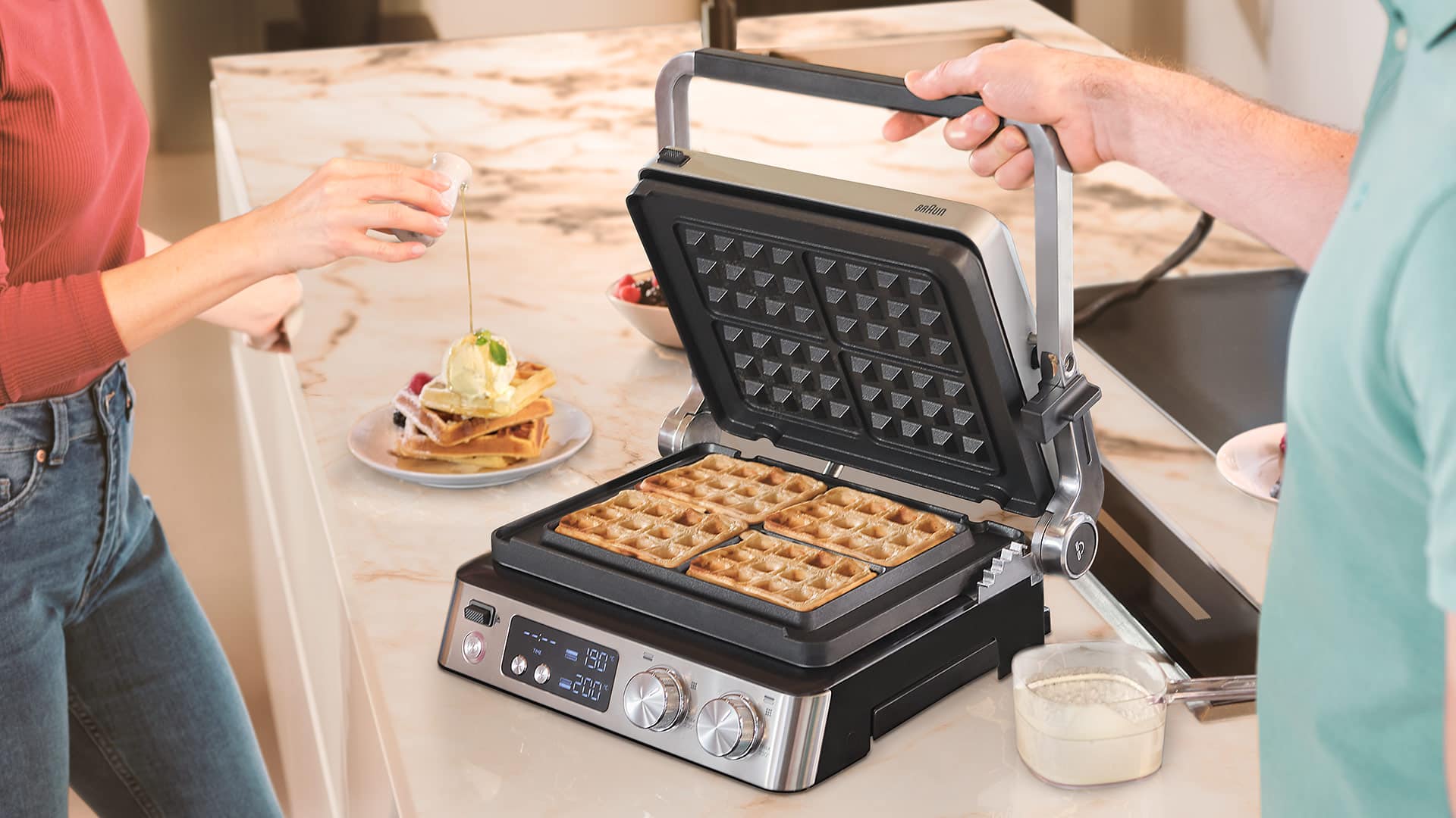 Person making waffles on a kitchen counter while another person adds syrup to plated waffles.