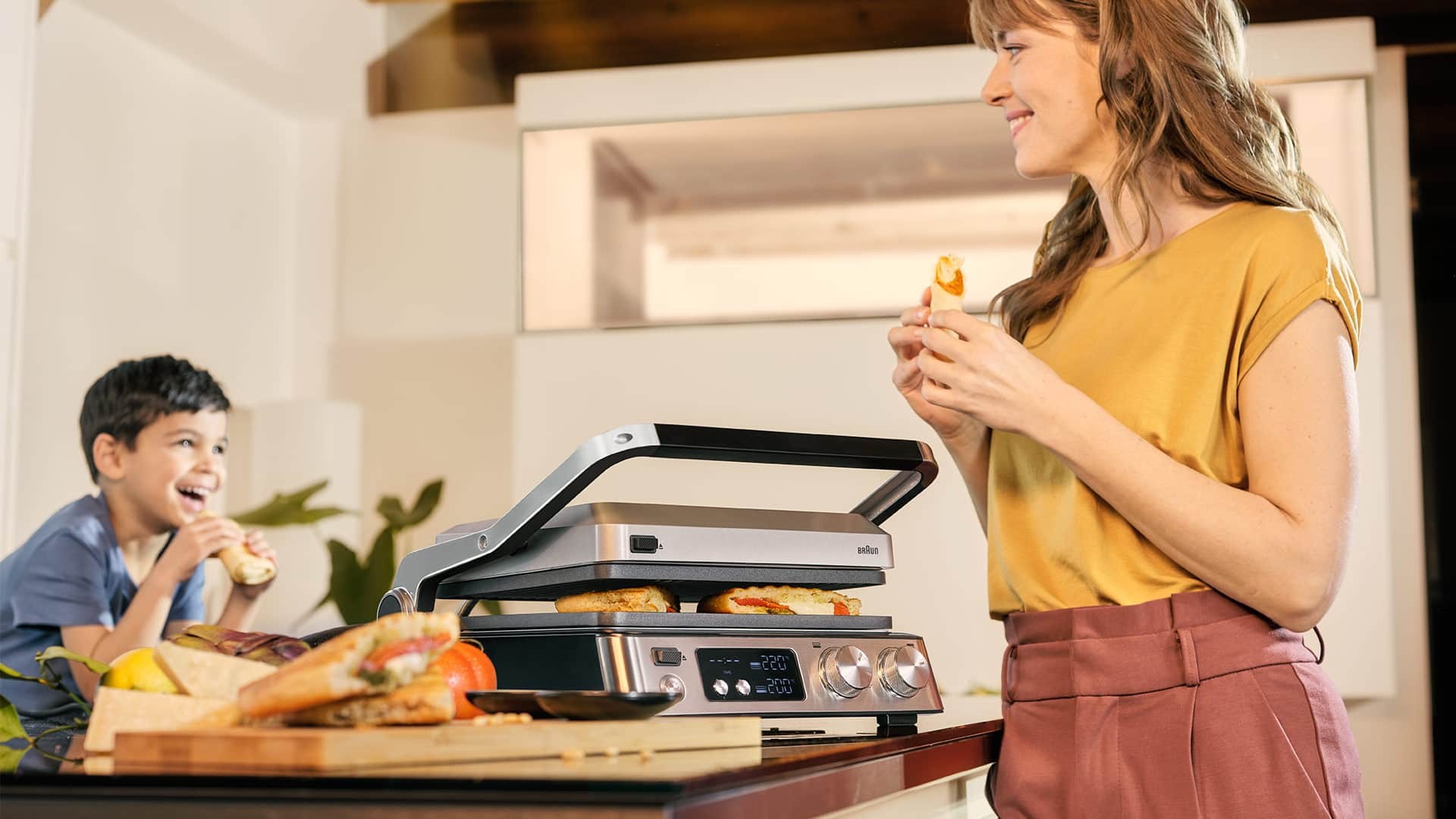 A woman and a boy enjoy slices of pizza together while seated in front of Braun's MultiGrill 7.